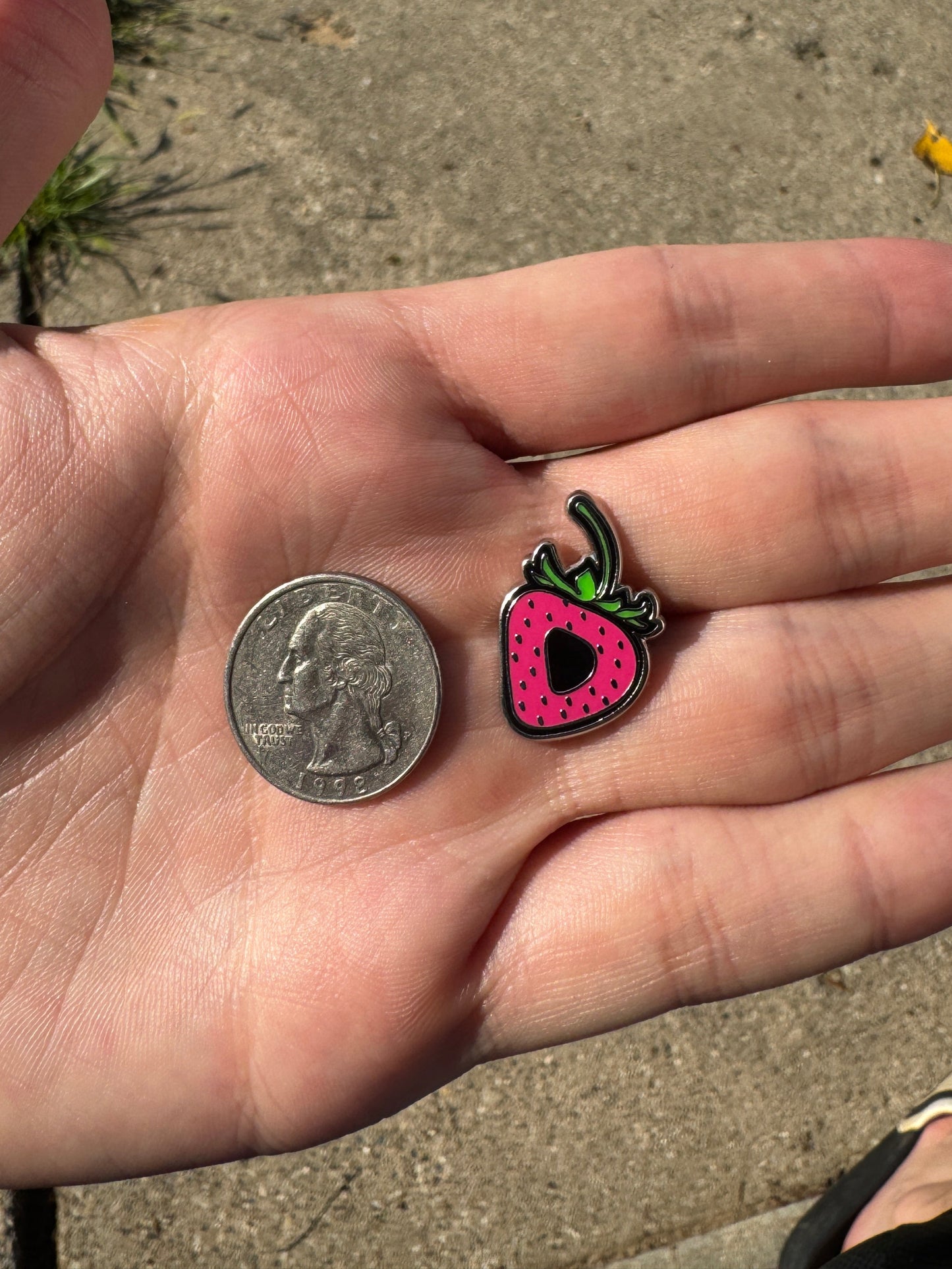 Hand holding a small colorful strawberry-shaped charm next to a coin on a concrete surface.