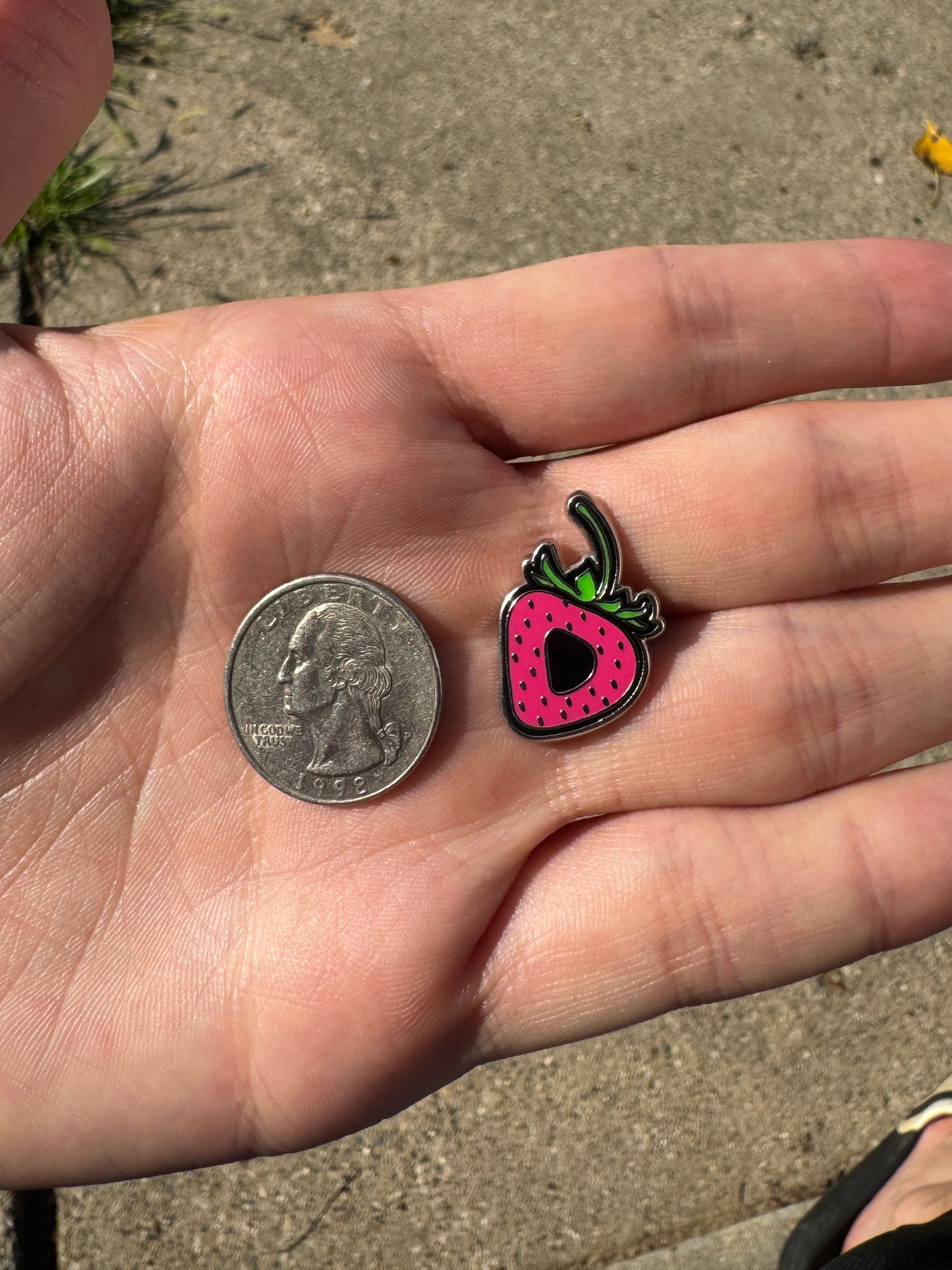 Hand holding a small colorful strawberry-shaped charm next to a coin on a concrete surface.