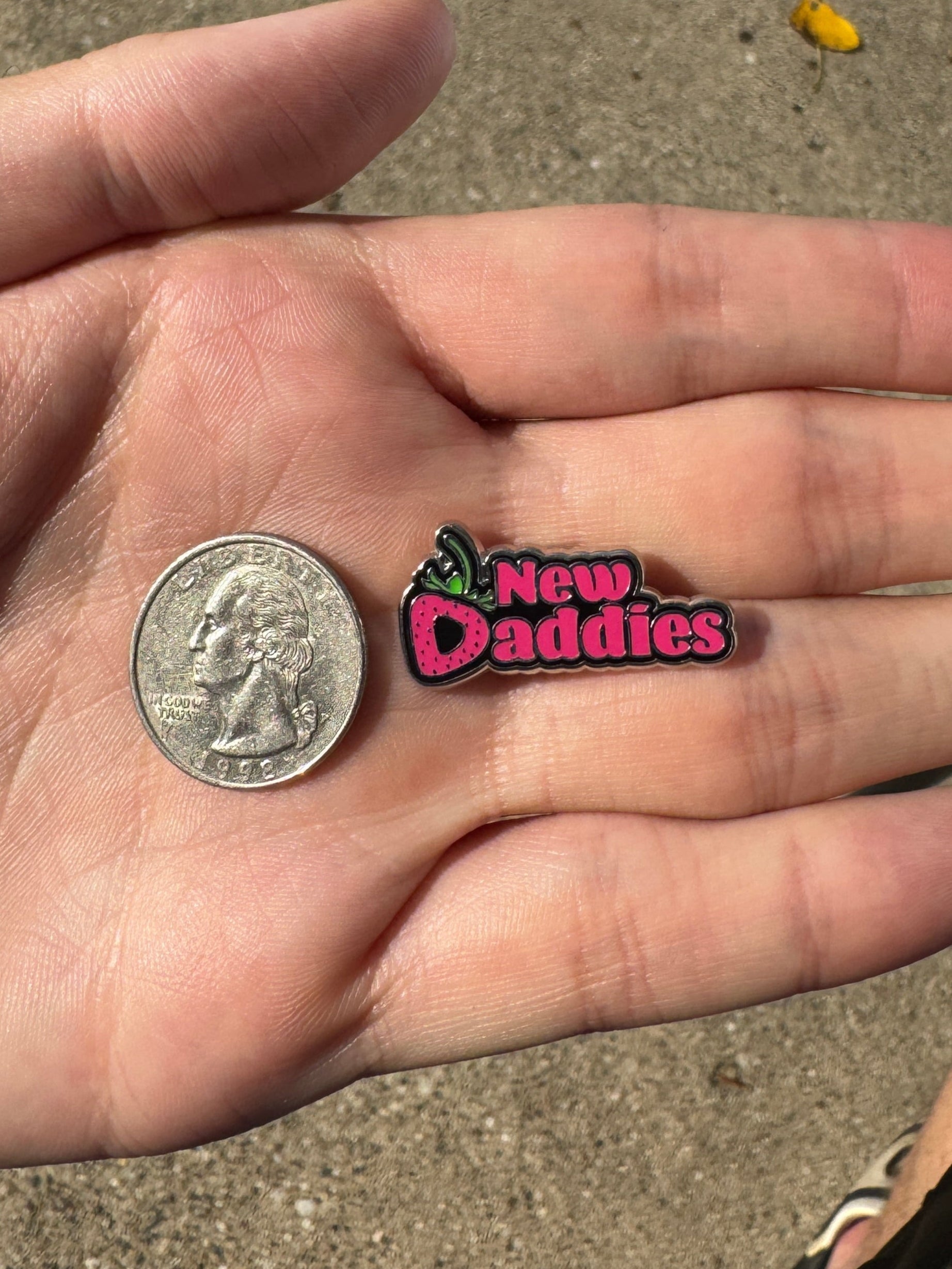 Small 'New Daddies' pin held in a hand with a coin for scale, on a concrete surface.